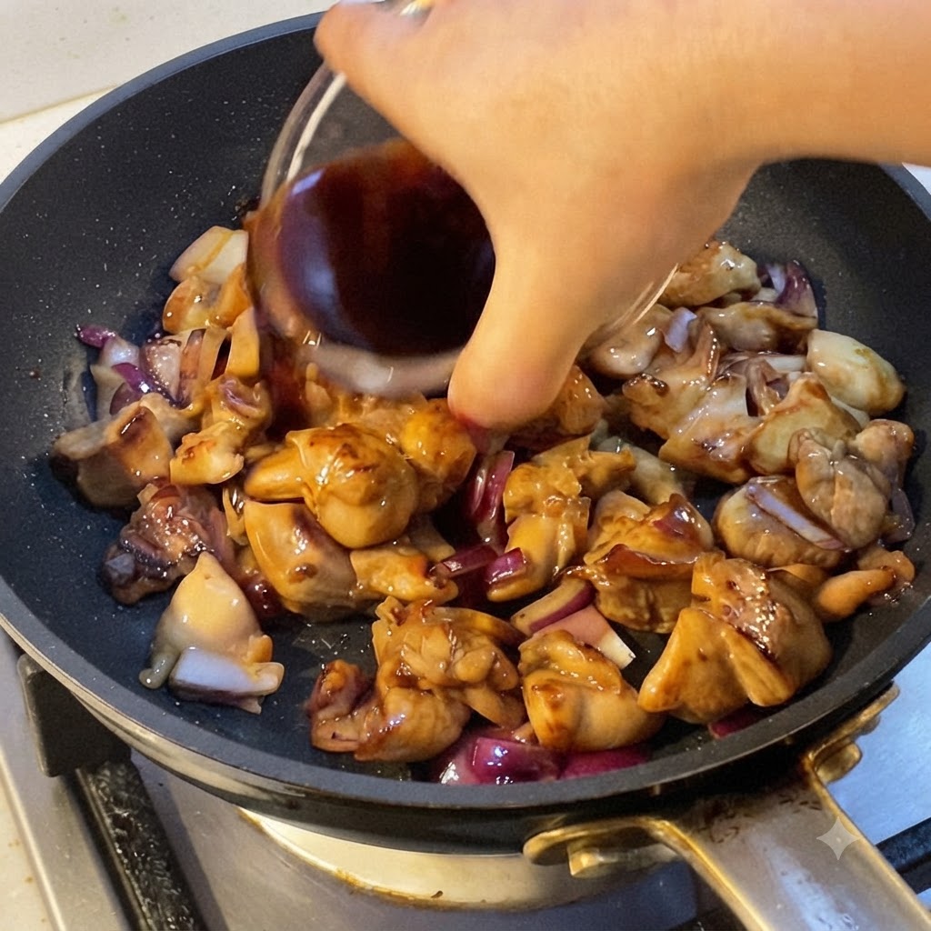 Eine Hand gießt dunkle Kochsoße in eine Pfanne mit gekochten Hähnchenstücken und roten Zwiebelstücken.