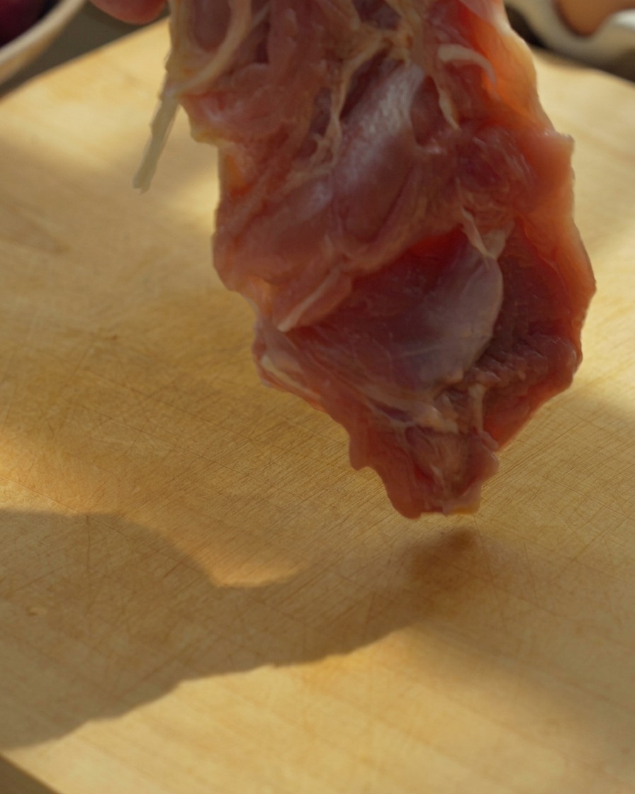 Entbeinte Hähnchenschenkelfilets werden auf einem Holzschneidebrett mit einem Messer in große Stücke geschnitten.