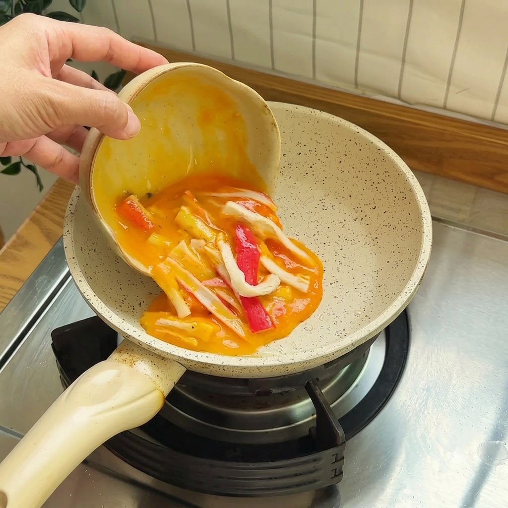 Verquirlte Eier vermischt mit roten und weißen Surimi-Stücken werden in eine kleine Antihaftpfanne gegossen.