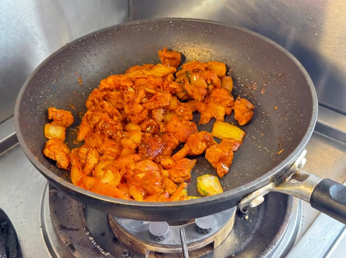Gewürfelte Hähnchenbrust, Zwiebeln und gehacktes Kimchi werden zusammen in einer dunklen Antihaft-Pfanne angebraten.