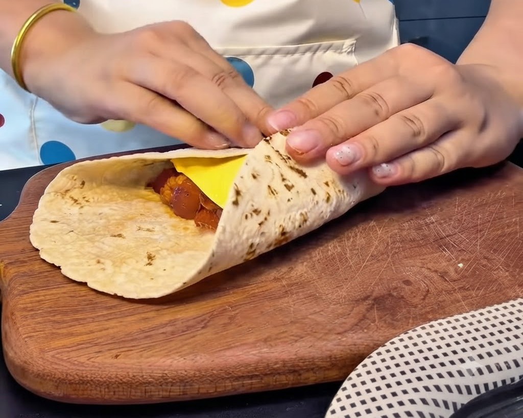 Hände falten die Ränder eines Fladenbrots fest über eine Scheibe Käse und die Füllung, um ein geschlossenes quadratisches Päckchen zu formen.