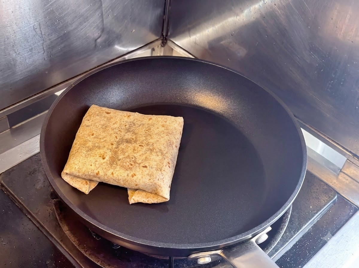 Ein gefalteter, quadratischer Fladenbrot-Wrap wird mit der Naht nach unten in einer trockenen schwarzen Antihaft-Pfanne geröstet.
