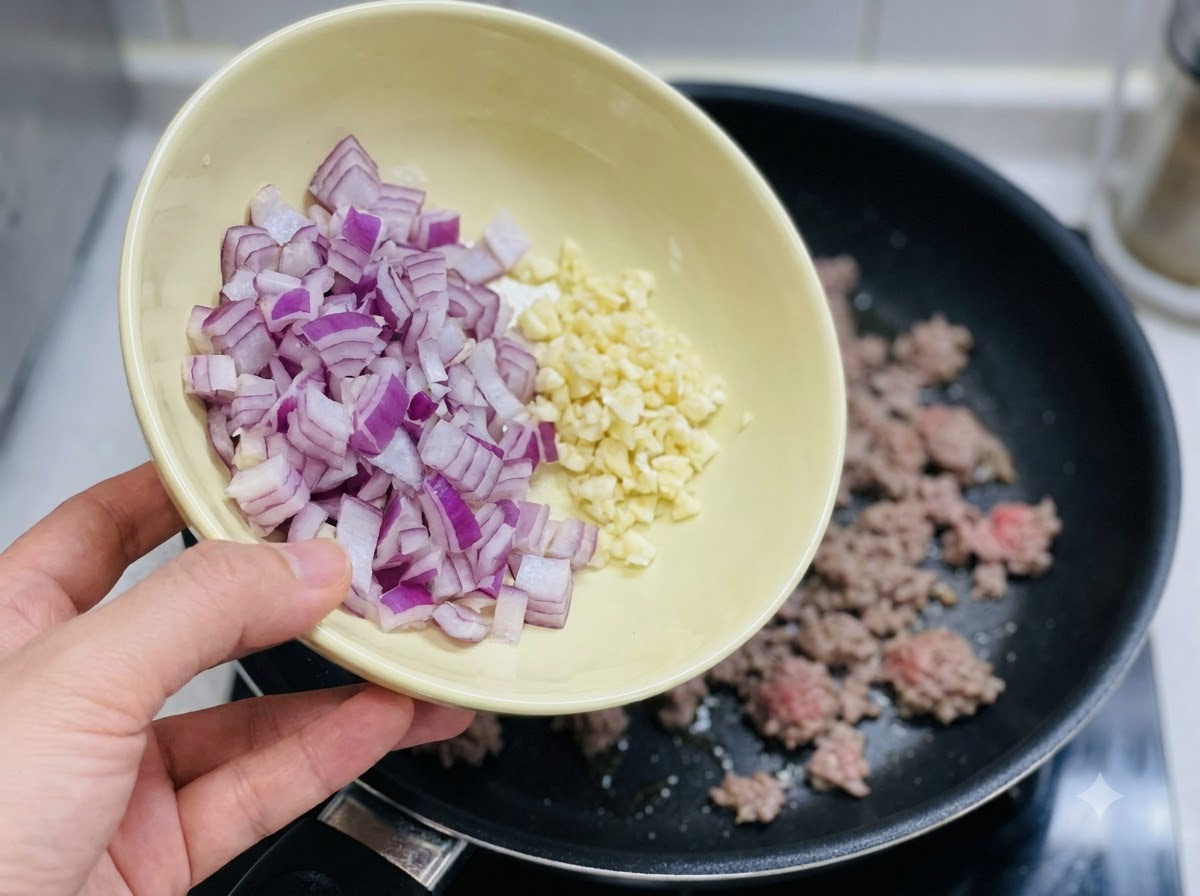 Eine Schüssel gehackte rote Zwiebel und gehackter Knoblauch wird in die Pfanne geschüttet.