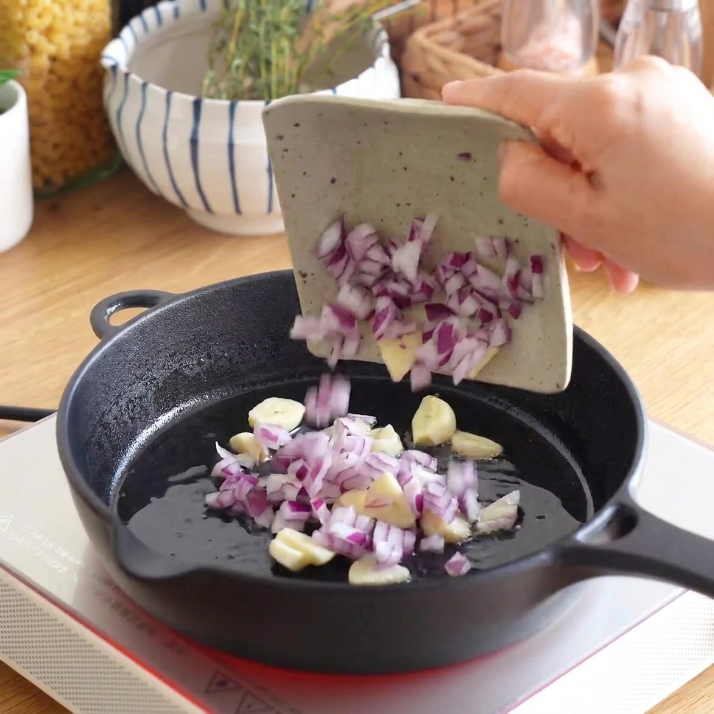 Gehackte rote Zwiebel und in Scheiben geschnittener Knoblauch werden aus einer kleinen Schale in eine heiße gusseiserne Pfanne gegeben.