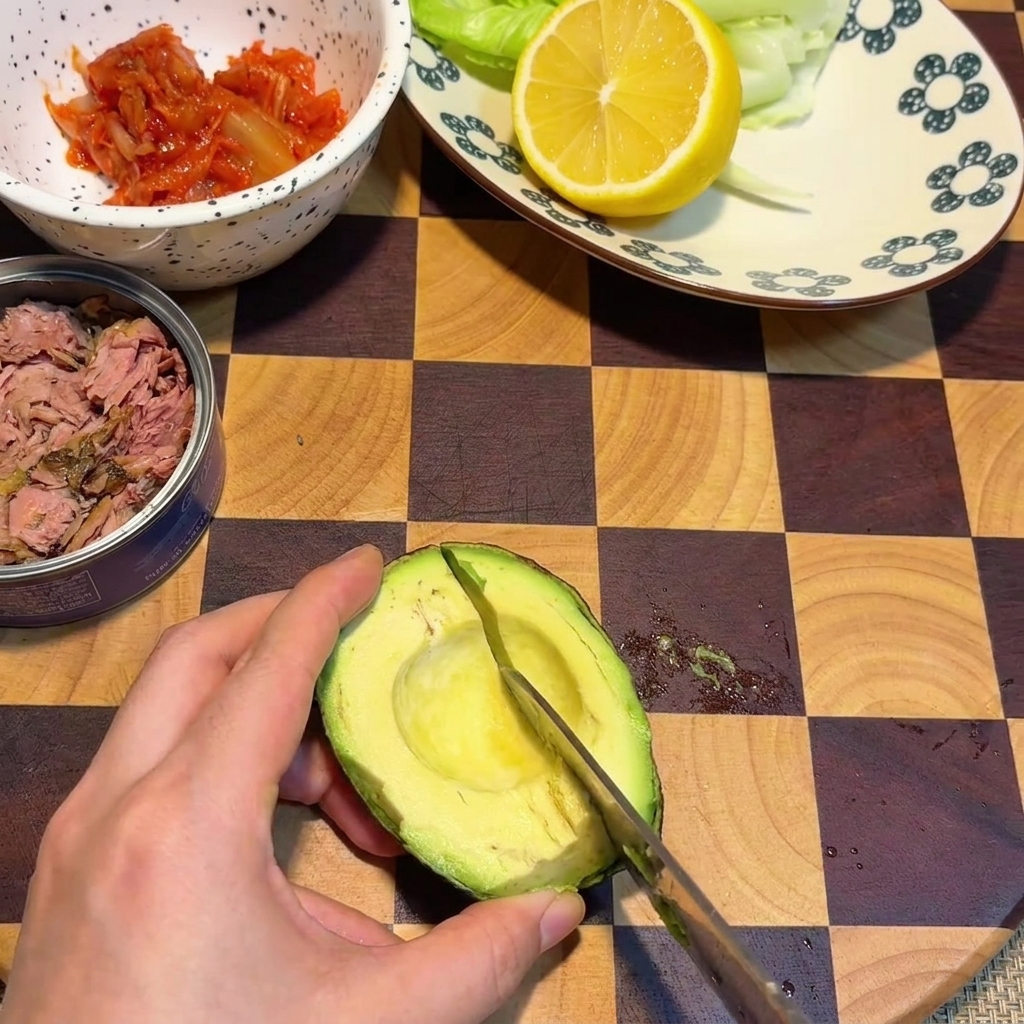 Eine Hand schneidet mit einem Messer karoförmige Würfel in eine Avocadahälfte auf einem karierten Holzschneidebrett.