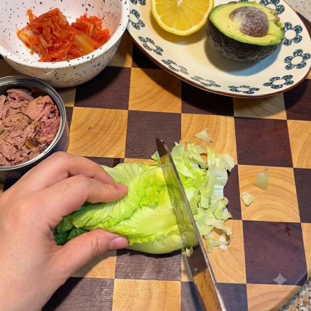 Ein Messer schneidet frische grüne Salatblätter auf einem karierten Schneidebrett neben einer Schüssel Kimchi und einer Dose Thunfisch.