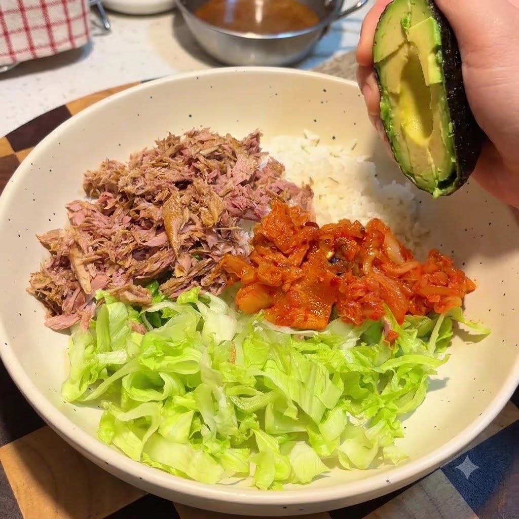 Eine Hand hält eine Avocadahälfte über eine große Schüssel mit Reis, Salat, Thunfisch und Kimchi.