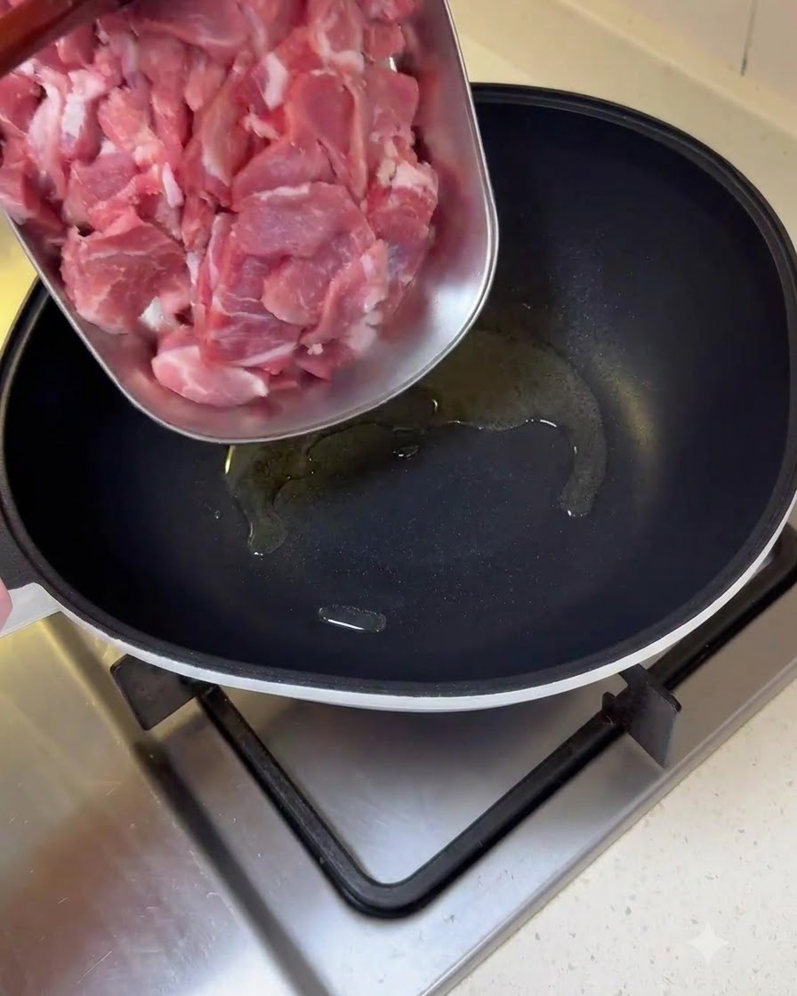 In Scheiben geschnittene rohe Schweinefleischstücke werden von einem Tablett in eine geölte Bratpfanne gegeben.