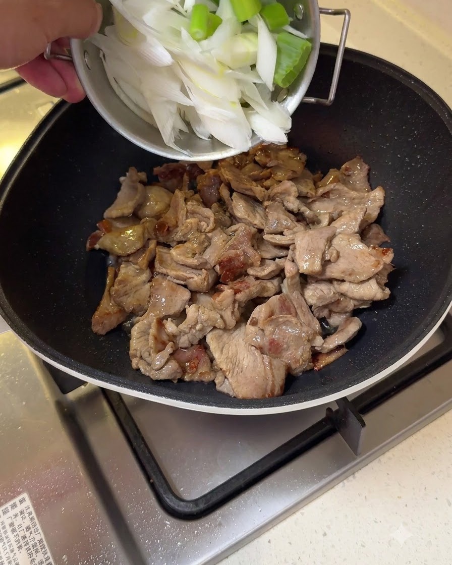 Gehackte große Frühlingszwiebelstücke werden zum angebratenen Schweinefleisch in einer Pfanne gegeben.
