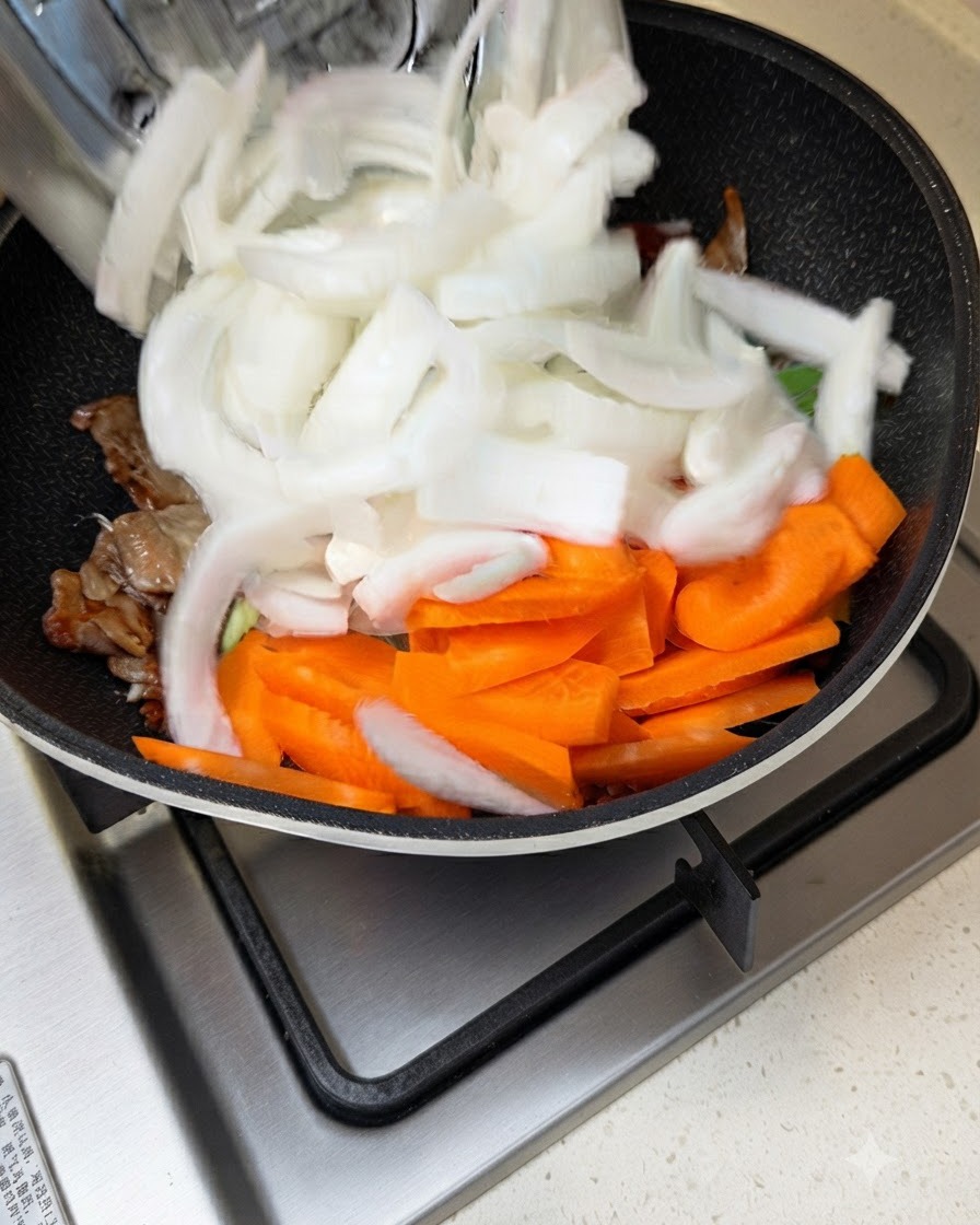 Geschnittene weiße Zwiebeln und Karotten werden in eine Pfanne mit anbratendem Fleisch und Sauce gegeben.