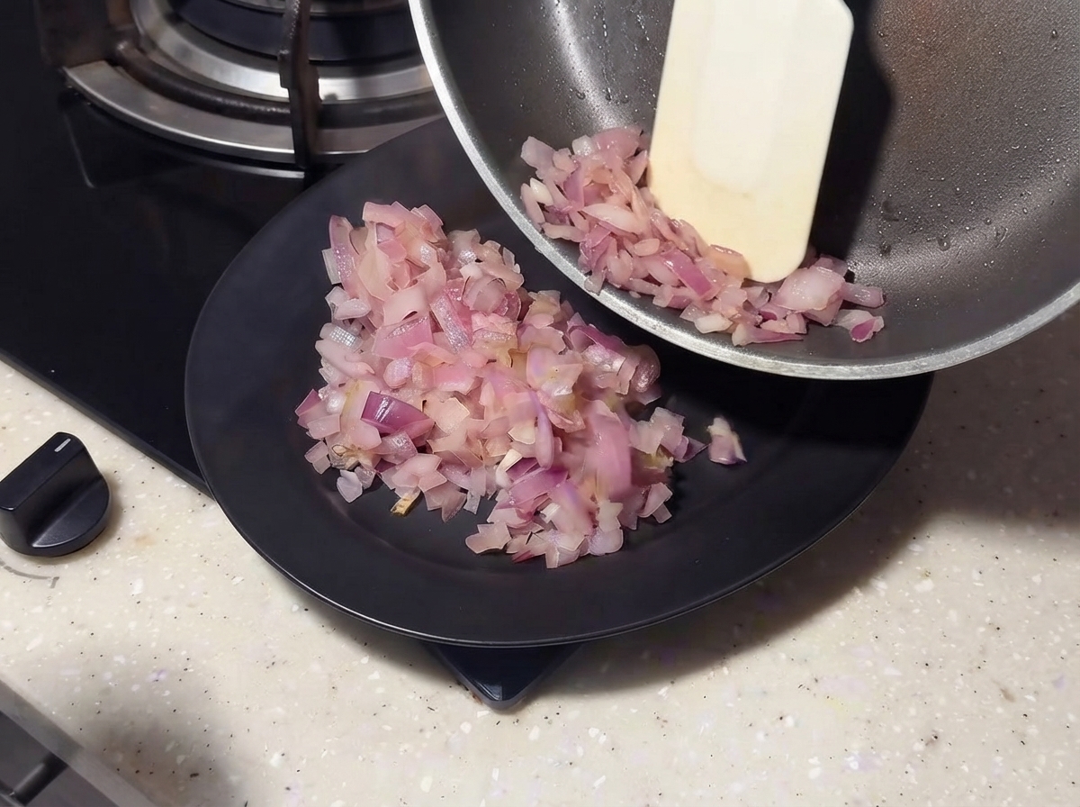 Angebratene Zwiebeln werden mit einem Spatel aus einer Pfanne auf einen schwarzen Teller geschoben.