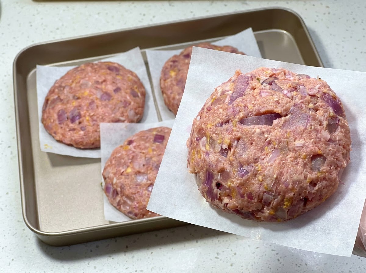 Ein frisch geformtes rohes Hamburger-Patty auf Backpapier, mit weiteren Patties auf einem Tablett dahinter.