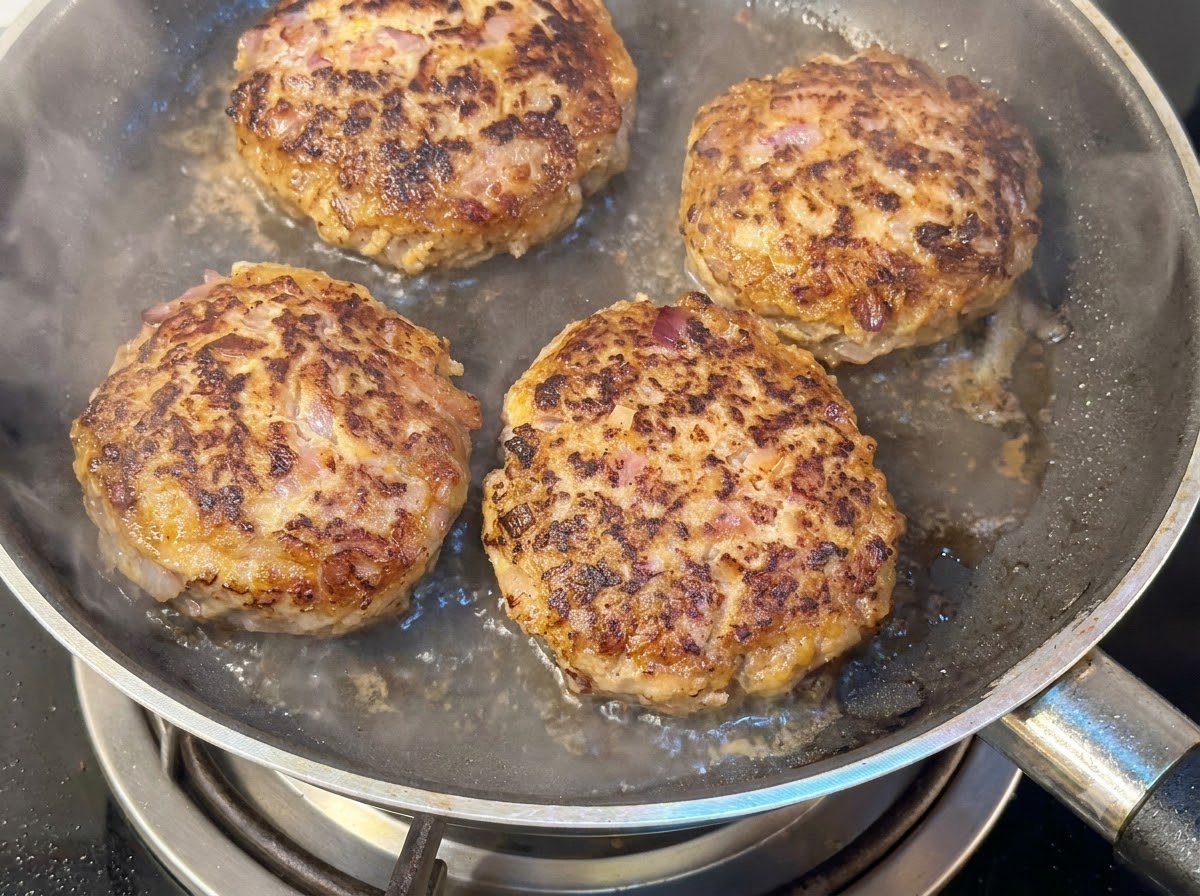 Vier dicke Hamburger-Patties braten in einer Pfanne mit Wasser und aufsteigendem Dampf unter einem Deckel.