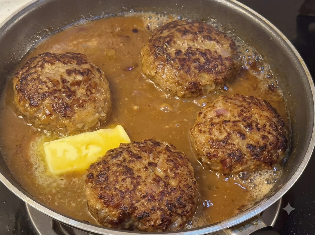 Hamburger-Patties köcheln in einer dicken braunen Sauce neben einem schmelzenden Butterstück.