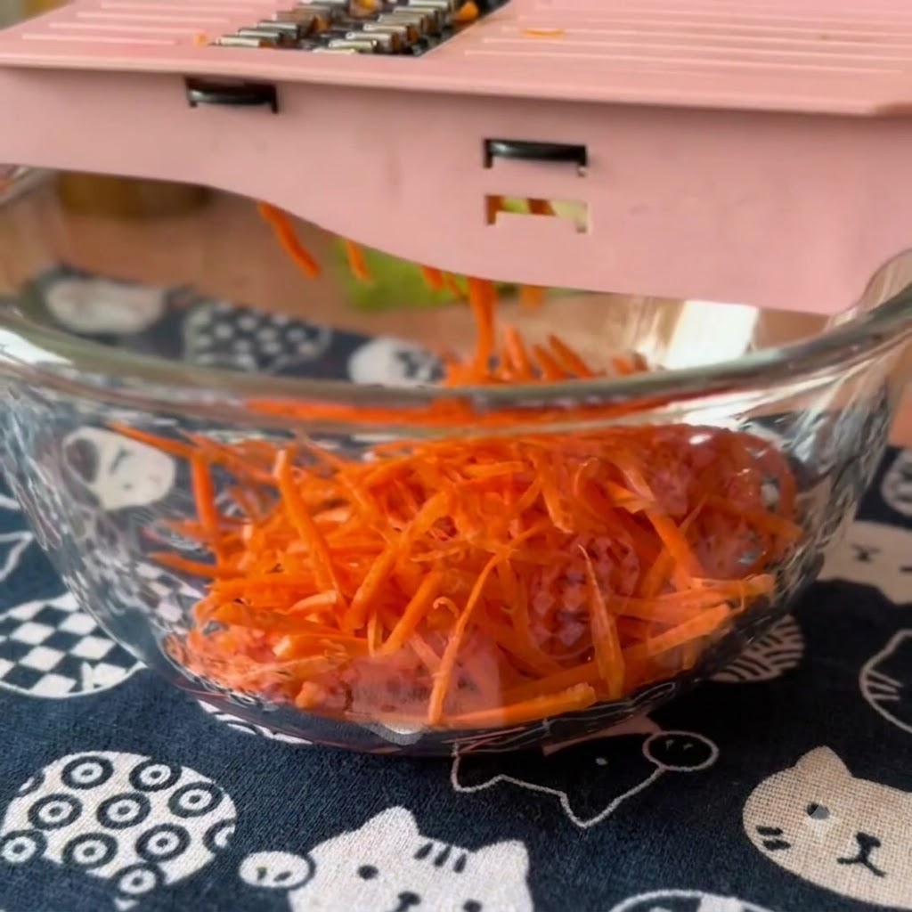 Frische Karotten und Gurken werden mit einem rosa Mandolinenschneider in eine Glasschüssel geraspelt.