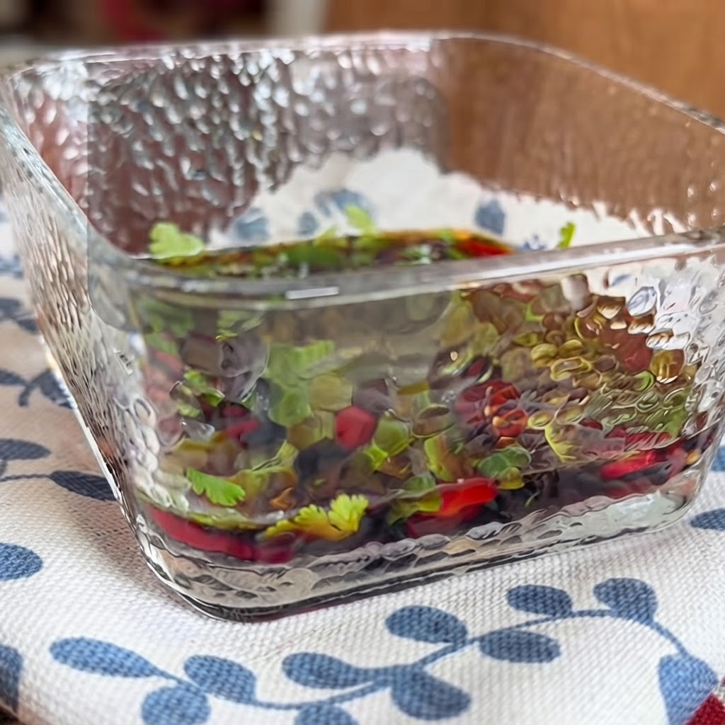 Nahaufnahme einer quadratischen Glasschüssel gefüllt mit einer herzhaften Dip-Sauce mit Kräutern und Chilischoten, die mit Stäbchen verrührt wird.