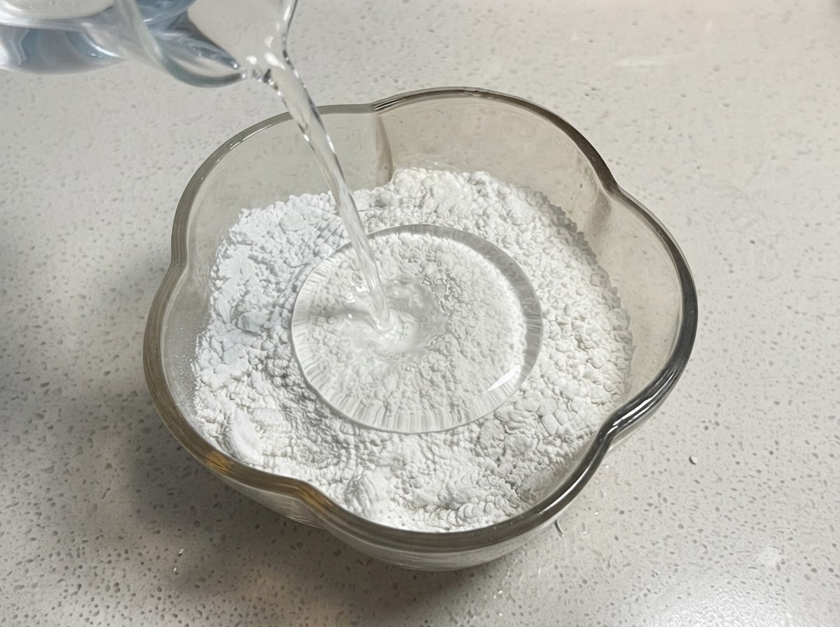 Wasser wird aus einem Messbecher in eine Glasschüssel mit Mehl und Stärke gegossen.