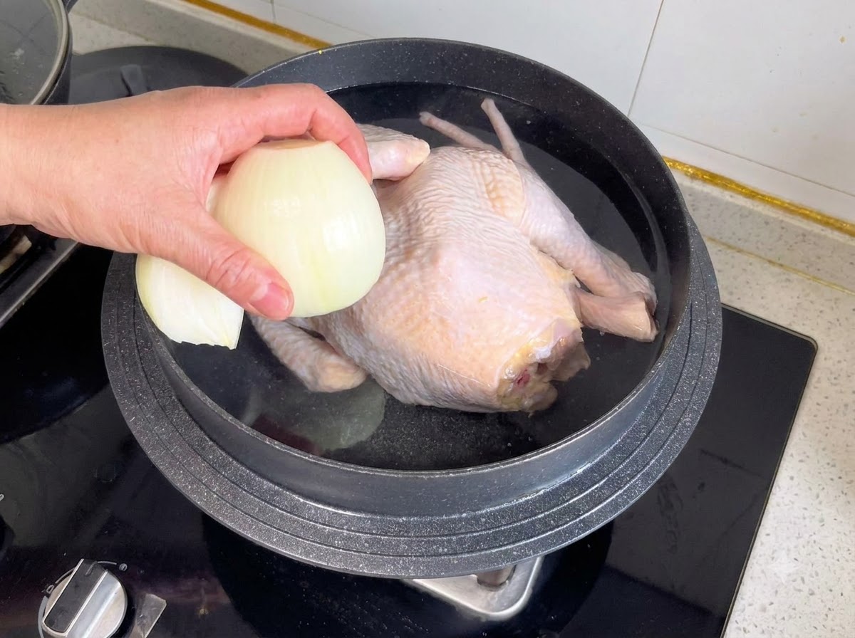 Eine halbe Zwiebel und andere Aromen werden in einen Topf mit einem ganzen Hähnchen und Wasser gegeben.