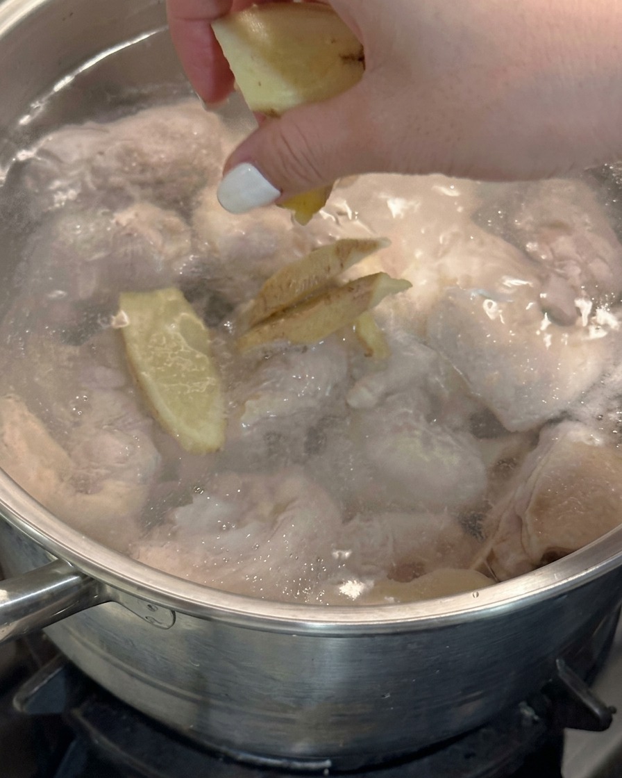 Eine Hand wirft frische Ingwerscheiben in einen Topf mit kochendem Wasser und Hähnchen.