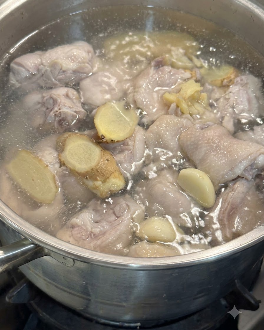 Knoblauchzehen und Ingwerscheiben schwimmen im Topf mit Hähnchenstücken in köchelndem Wasser.