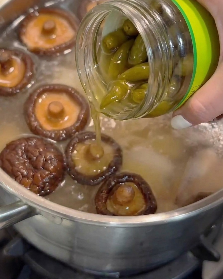 Eingelegter Saft von geschälten Chilischoten wird aus einem Glas in einen Topf mit köchelnder Suppe mit Shiitake-Pilzen gegossen.