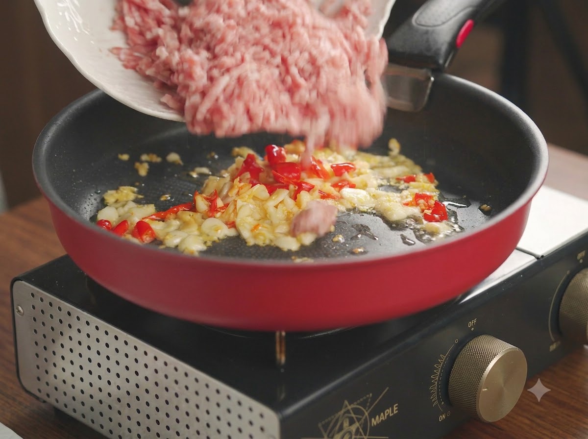 Rohes Schweinehackfleisch wird von einem weißen Teller in eine rote Pfanne mit gebratenem Knoblauch und Chili gefüllt.