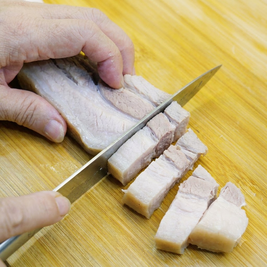 Blanchierter Schweinebauch wird auf einem Holzschneidebrett in mundgerechte Stücke geschnitten.