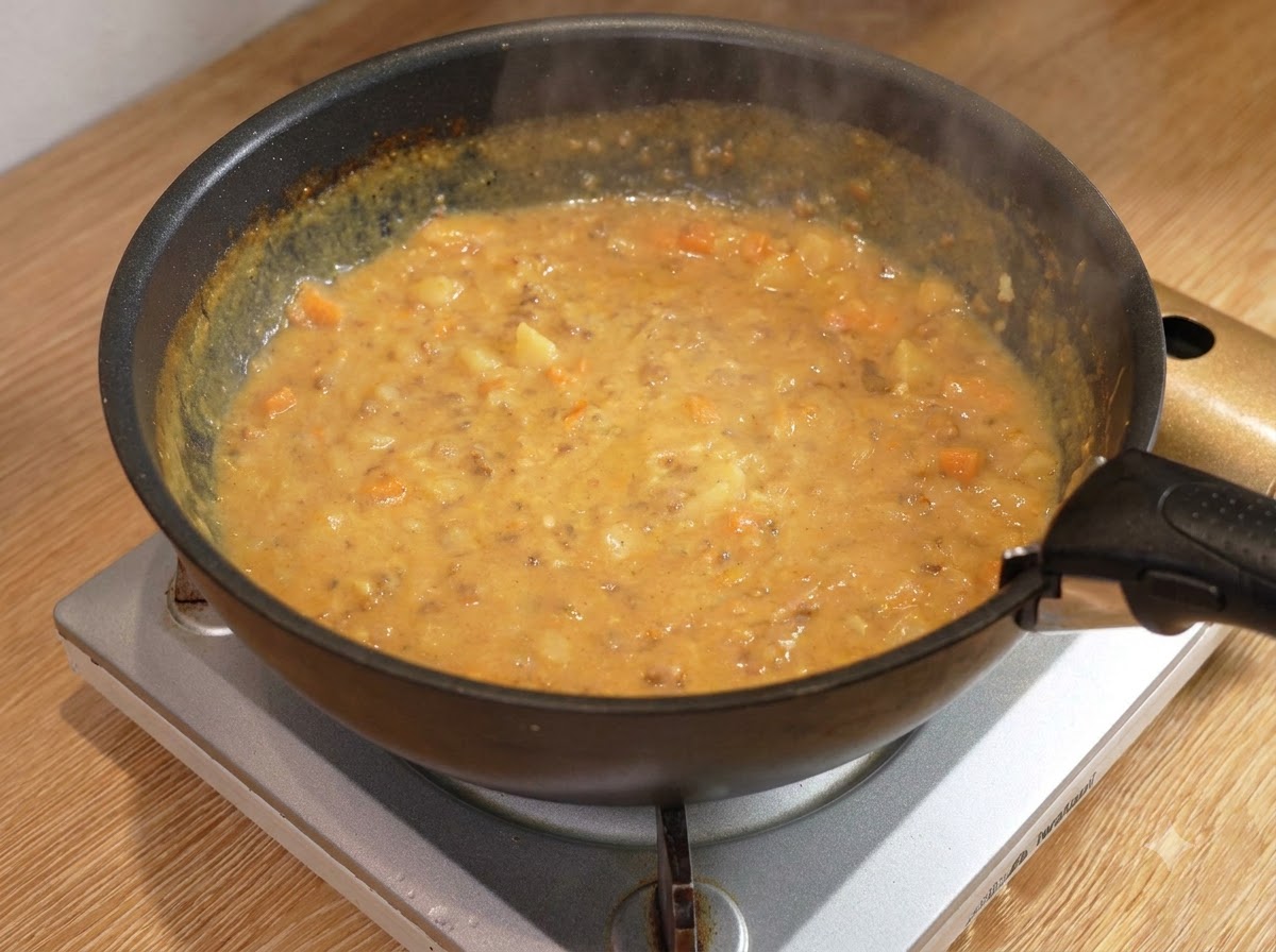 Ein Holzlöffel rührt eine dicke, blubbernde Curry-Hackfleischsoße in einer schwarzen Pfanne auf einer Kochstelle um.