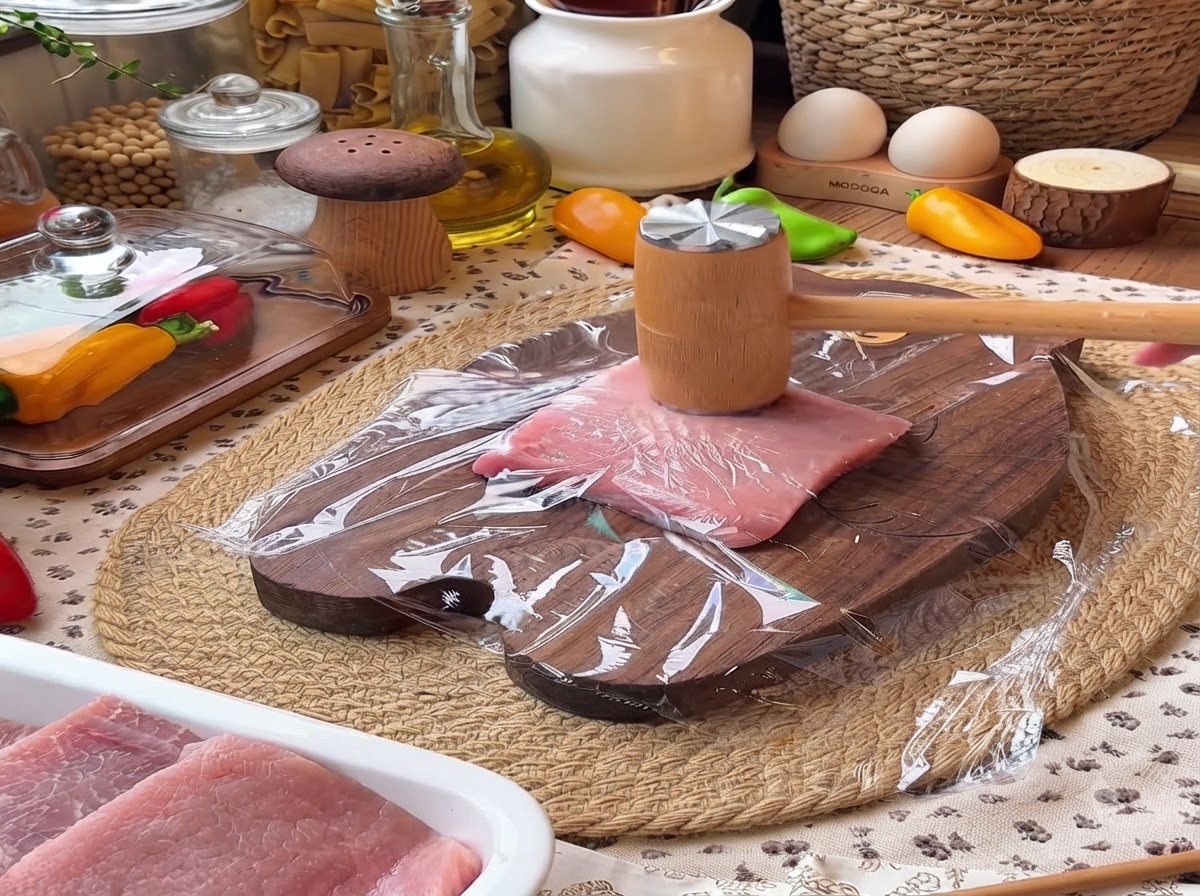 Ein Fleischklopfer aus Holz klopft ein mit Frischhaltefolie bedecktes Stück Schweinefilet auf einem Holzschneidebrett flach.