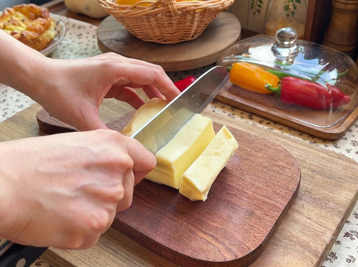 Ein Paar Hände schneidet mit einem Messer einen Block Mozzarellakäse auf einem Holzschneidebrett in kleine rechteckige Stücke.