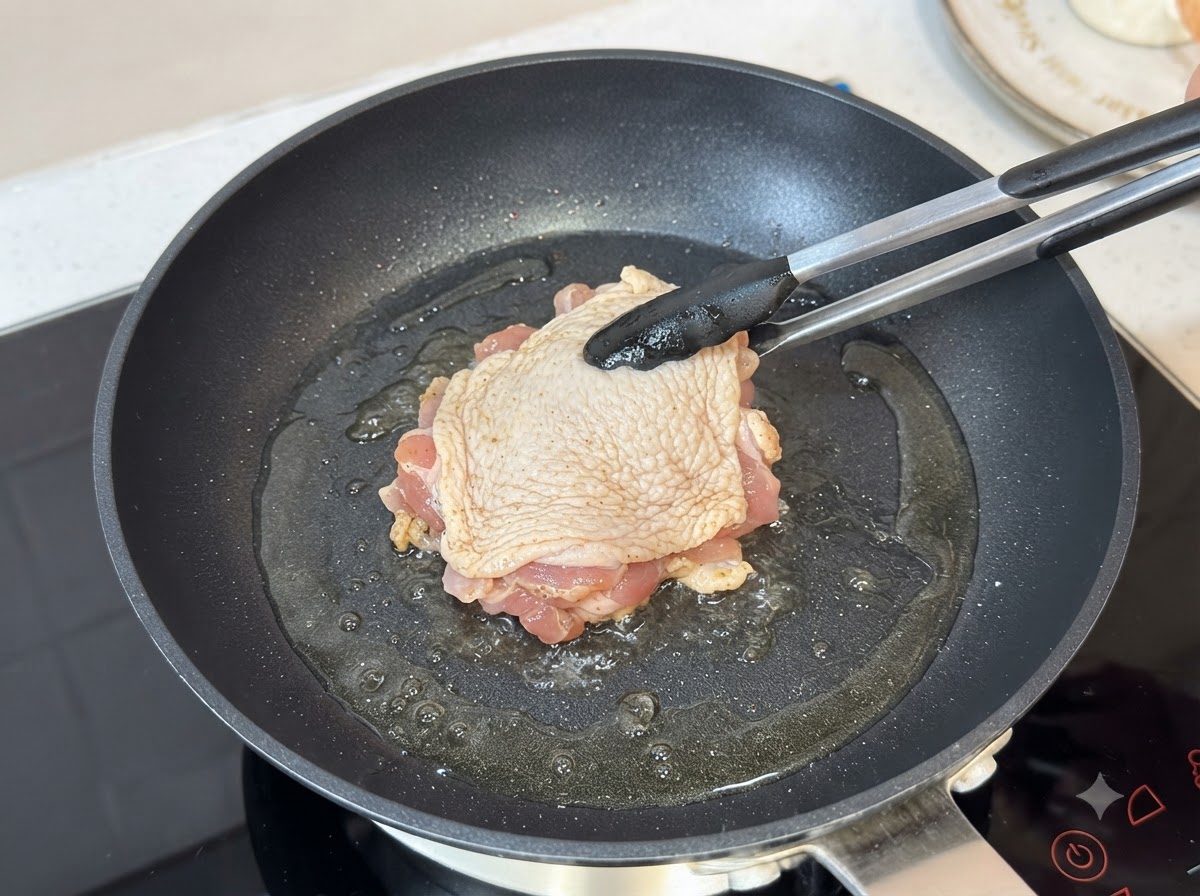 Ein gewürzter Hähnchenschenkel brät mit der Hautseite nach unten in einer heißen, geölten Pfanne unter Verwendung einer Zange.