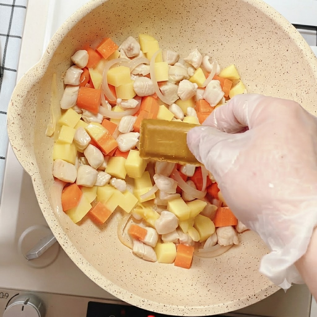 Eine Hand legt einen Currywürfel in die mit gemischtem Gemüse und Hähnchen gefüllte Pfanne.