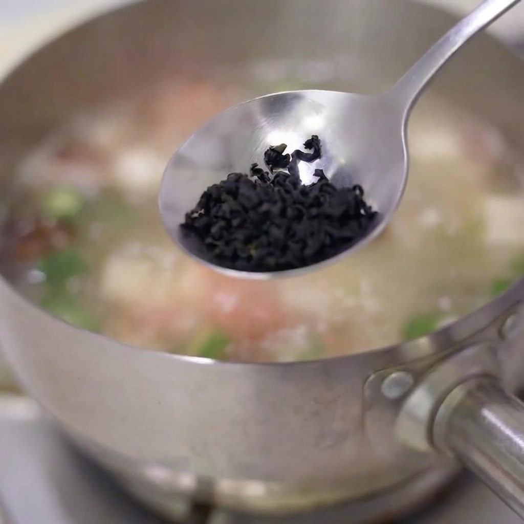 Ein Löffel getrockneter Algenflocken wird in einen köchelnden Edelstahltopf mit Suppe gegeben.