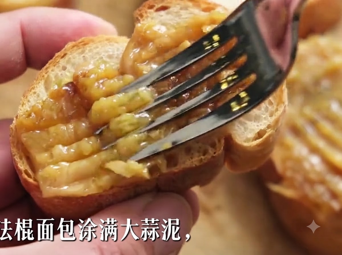 Eine Gabel streicht dicke geröstete Knoblauchpaste auf eine Scheibe knuspriges Baguette.