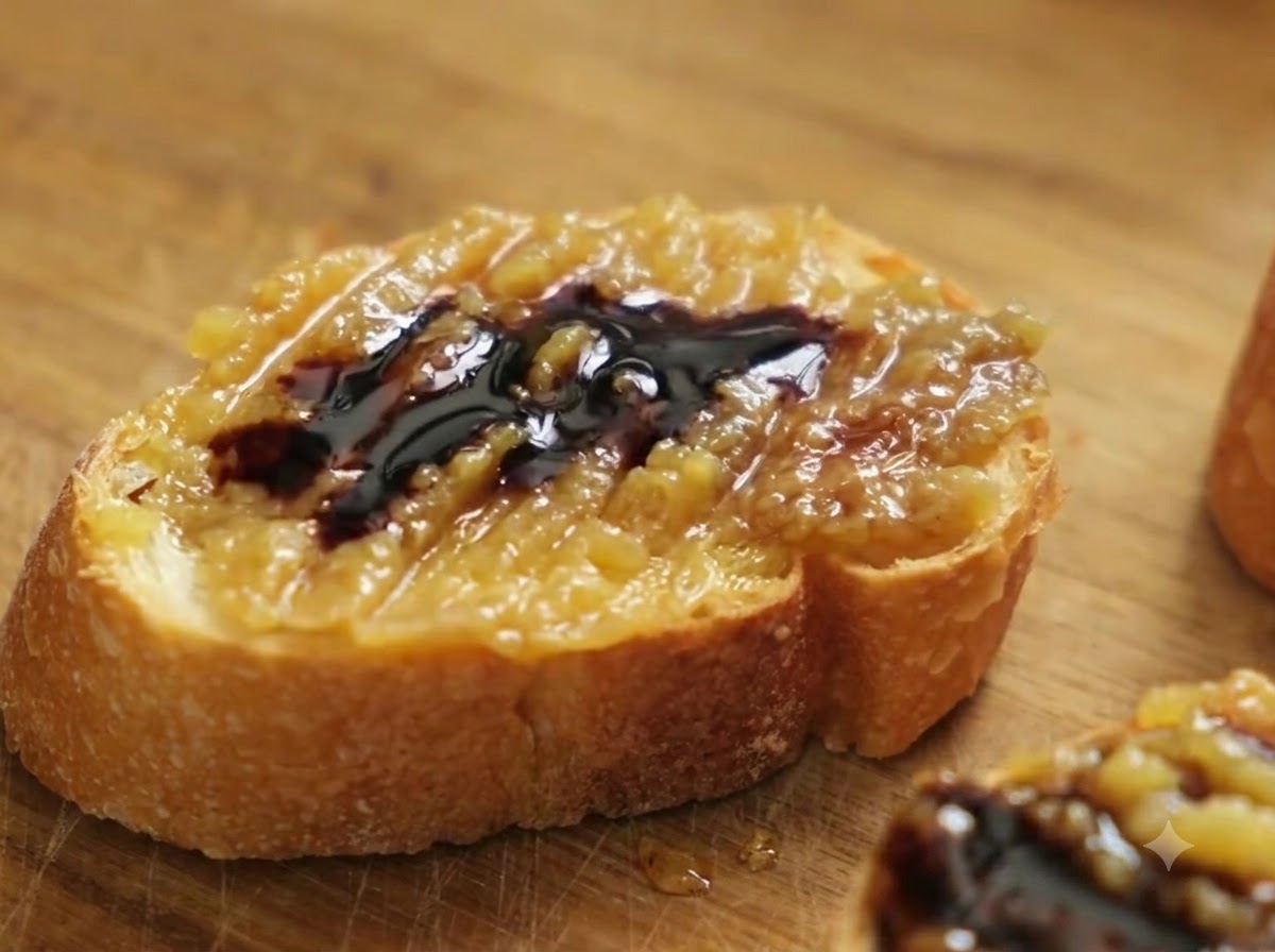 Nahaufnahme einer Baguettescheibe mit Knoblauch-Confit-Paste und Balsamico, auf die grobes Meersalz gestreut wird.