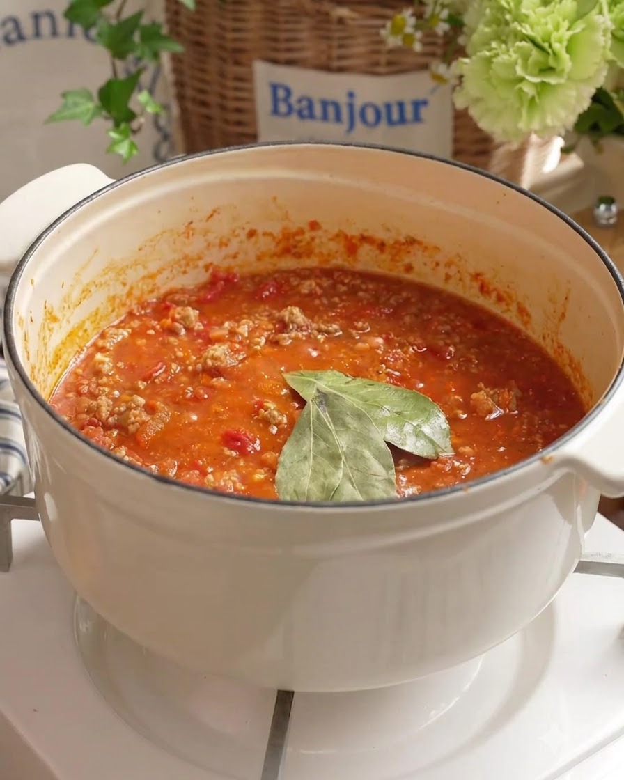 Lorbeerblätter und frisches Wasser werden in die köchelnde Tomaten-Hackfleischsoße in einem weißen Topf gegeben.