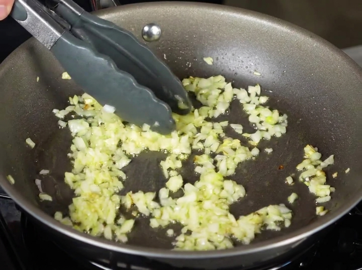 Gekochte Tagliatelle-Nudeln werden mit einer Zange in eine Pfanne mit gehackten, angebratenen Zwiebeln gegeben.