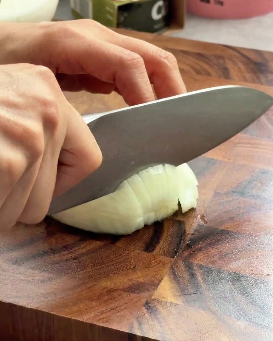 Ein scharfes Kochmesser schneidet auf einem Holzbrett durch eine weiße Zwiebel.