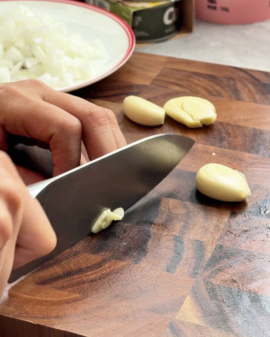 Knoblauchzehen werden mit einem Kochmesser auf einer Holzoberfläche fein gehackt.