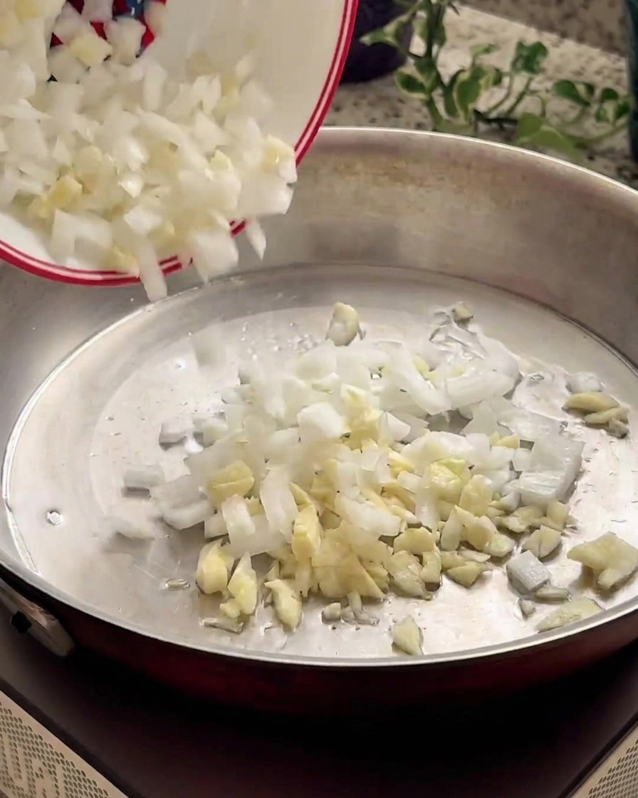 Gehackte Zwiebeln und Knoblauch gleiten aus einer kleinen weißen Schüssel in eine warme Metallpfanne.