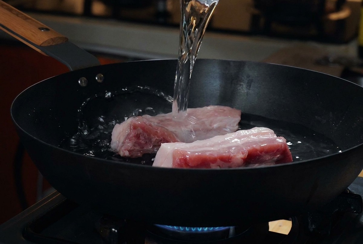 Dicke Stücke roher Schweinebauch liegen in einer kalten schwarzen Pfanne, während Wasser aus einem Messbecher darübergegossen wird.