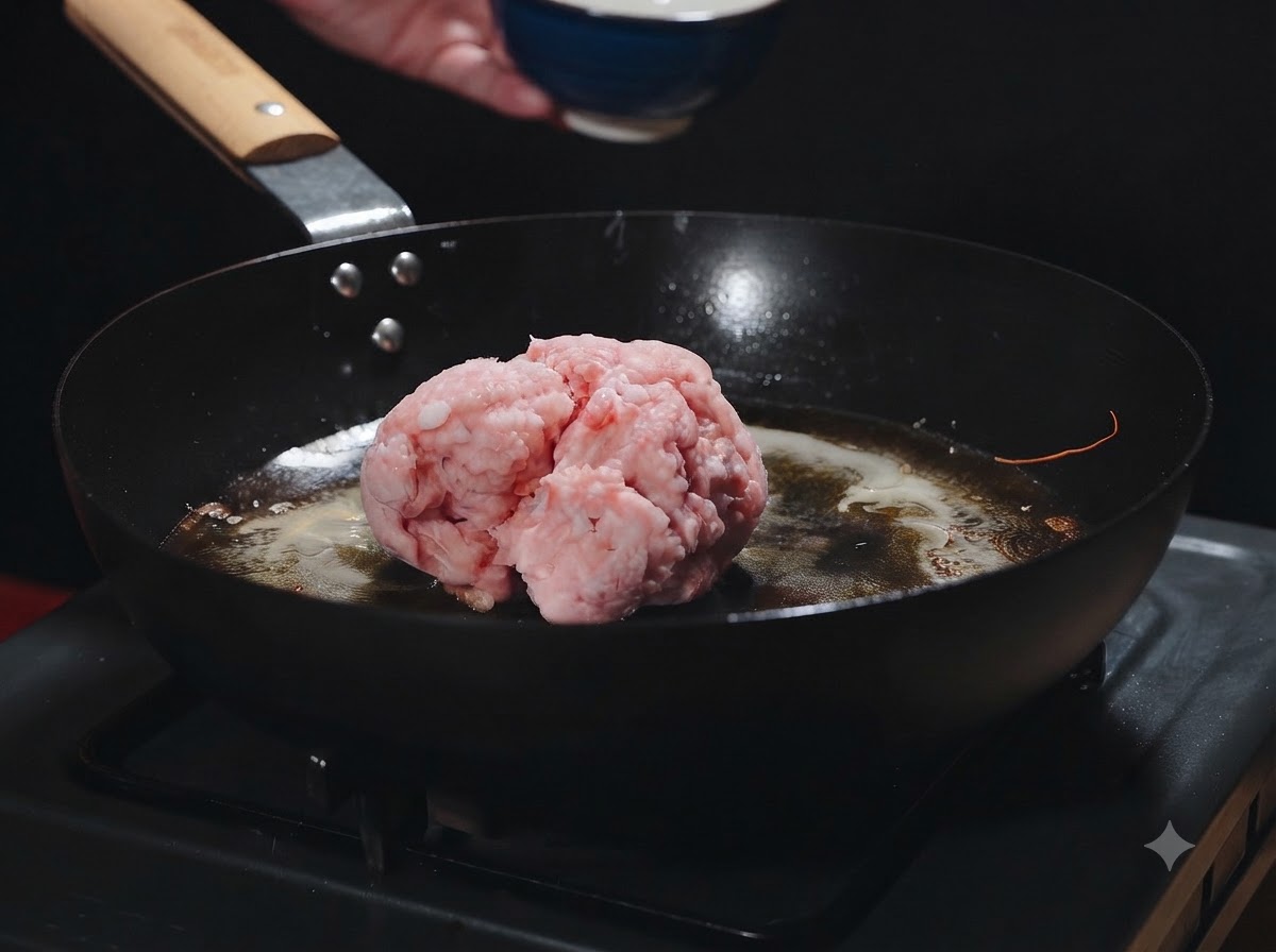 Eine Kugel rohes Hackfleisch liegt in einer heißen Pfanne mit Garnelenöl, während sie hinzugefügt wird.