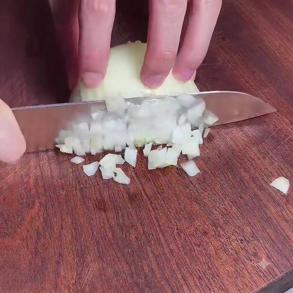 Hände halten ein Messer und würfeln eine Zwiebel fein auf einem Holzschneidebrett.