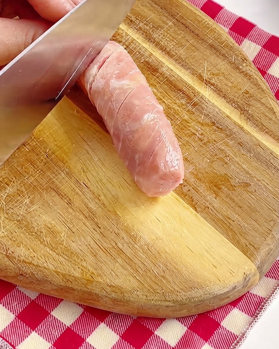 Eine Hand schneidet die Oberfläche eines ganzen rosa Würstchens mit einem Messer auf einem Holzbrett vorsichtig ein.