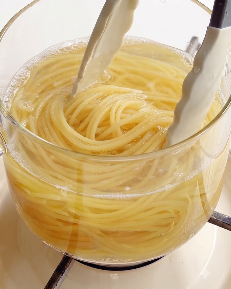 Mit einer weißen Küchenzange werden perfekt gekochte Spaghetti aus einem Glastopf mit kochendem Wasser gehoben.
