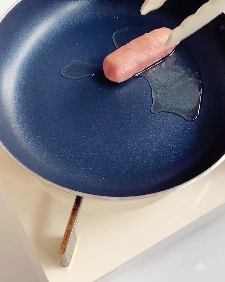 Eingeschnittene Würstchen braten in einer dunklen beschichteten Pfanne mit etwas Öl.