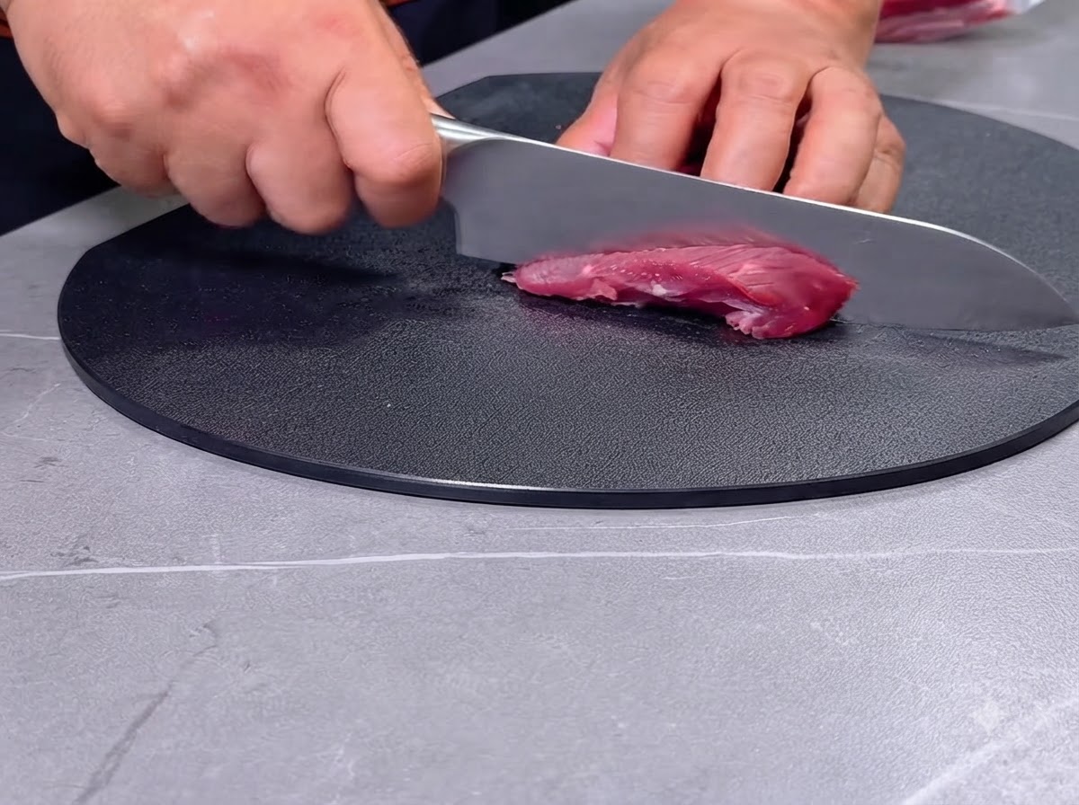 Eine Person schneidet rohes rotes Fleisch mit einem Messer auf einem schwarzen Schneidebrett in Stücke.