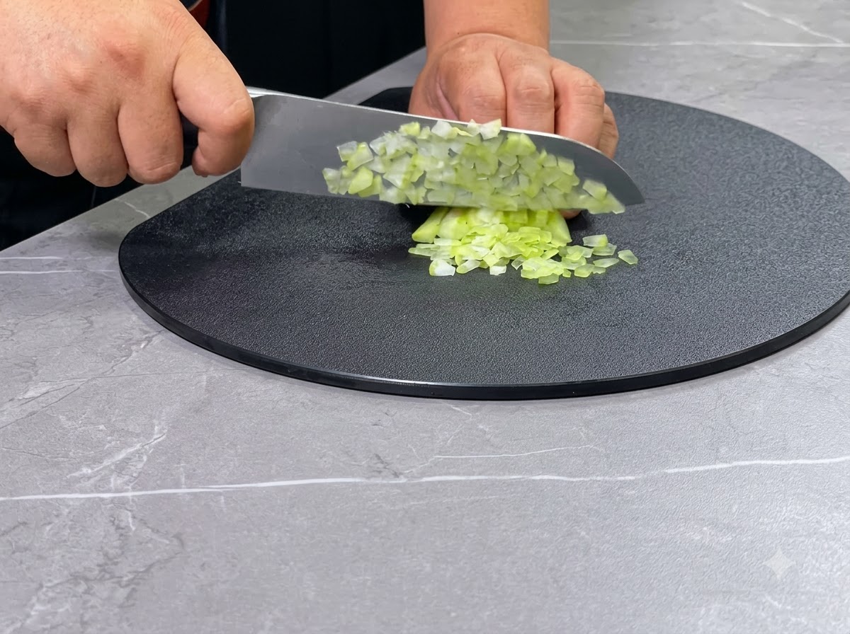 Zwei Hände würfeln grüne Selleriestangen mit einem Kochmesser auf einem schwarzen, runden Schneidebrett.
