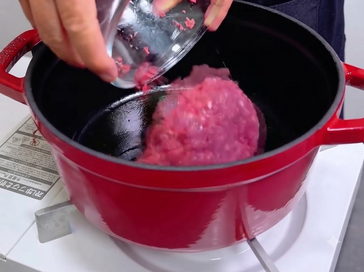 Eine Schüssel gießt rohes, zerkleinertes Fleisch in einen heißen, roten Gusseisentopf.