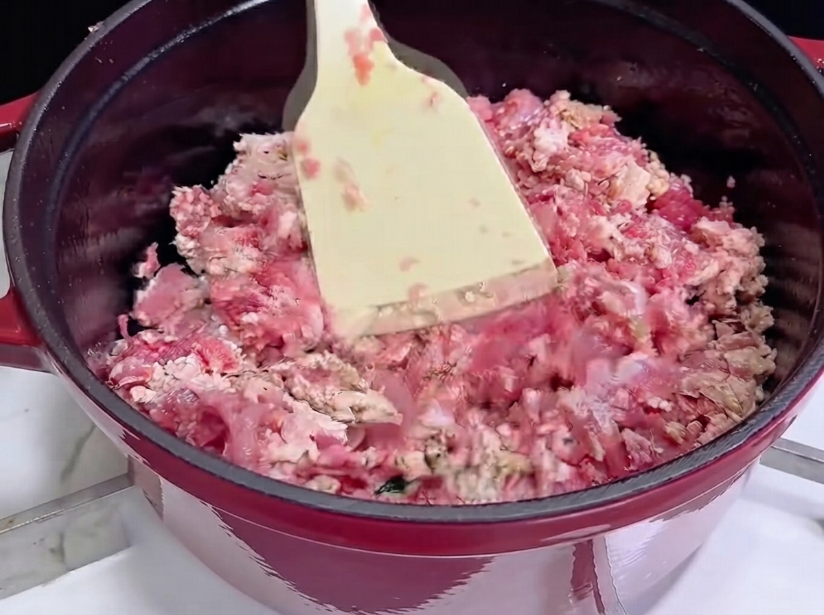 Anbraten von Hackfleisch mit einem weißen Spatel in einem roten Gusseisentopf, während es anfängt, braun zu werden.